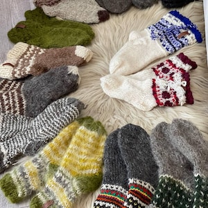 Puede incluir: Una colección de calcetines y mitones tejidos a mano en varios colores y patrones. Los calcetines presentan diseños en blanco, verde, marrón, gris y amarillo. Los mitones son principalmente grises con acentos coloridos. Los artículos están dispuestos sobre una superficie blanca y esponjosa.