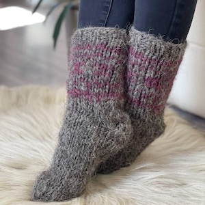 May include: Chunky grey knitted socks with a pink stripe design. These thick socks look warm and are ideal for colder weather. The socks are displayed on a white fluffy rug.