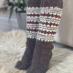 May include: Knee-high knit socks in shades of brown, white, and red. The socks feature a patterned design with a dark brown cuff and foot, and a white and red pattern on the leg. The socks are made of a thick, warm material.