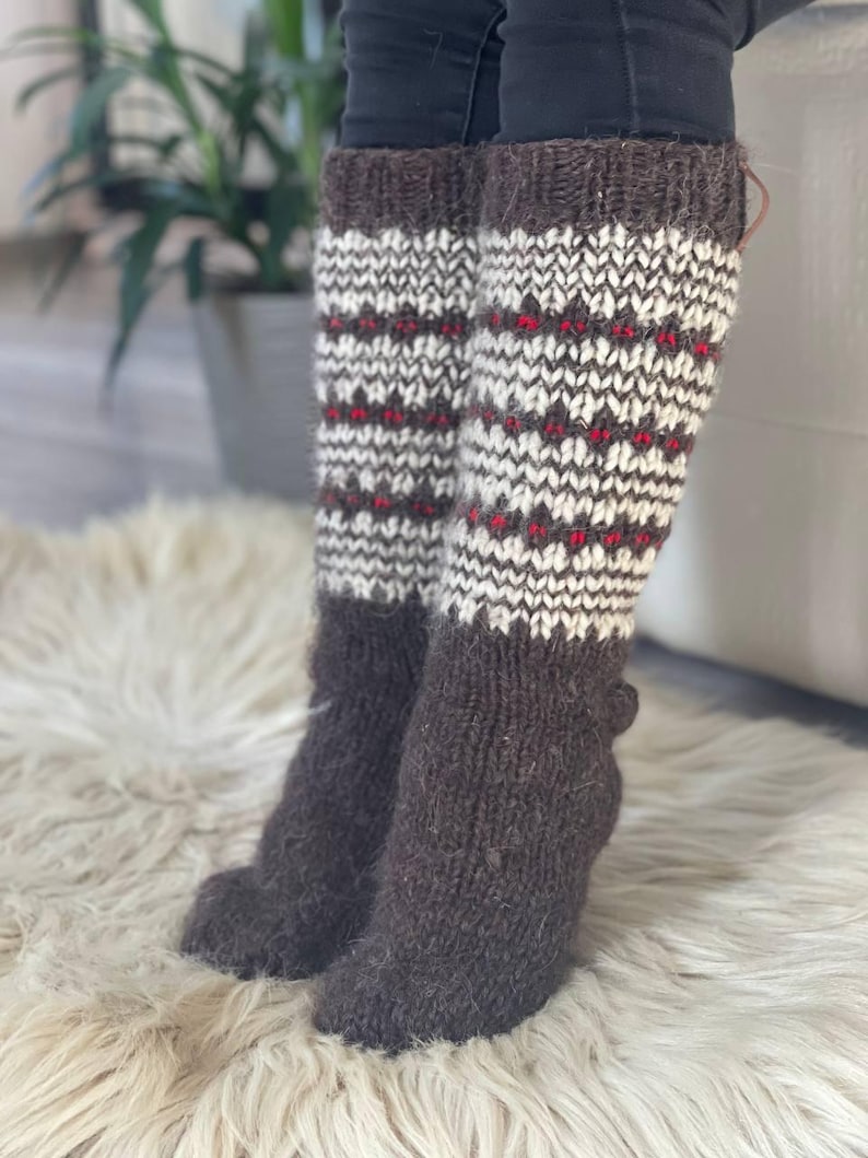 May include: Knee-high knit socks in shades of brown, white, and red. The socks feature a patterned design with a dark brown cuff and foot, and a white and red pattern on the leg. The socks are made of a thick, warm material.