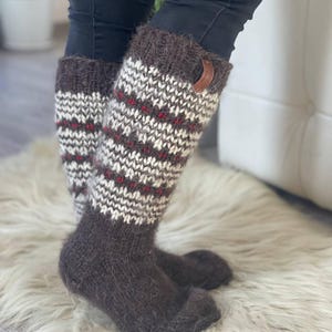 May include: Knee-high knit socks in brown, white, and red. The socks have a textured pattern with a brown cuff and a small leather tag. The socks are worn with black pants and are on a white fur rug.