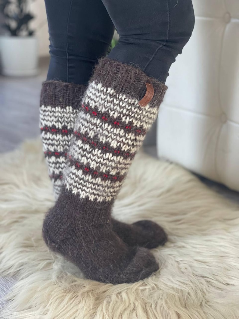 May include: Knee-high knit socks in brown, white, and red. The socks have a textured pattern with a brown cuff and a small leather tag. The socks are worn with black pants and are on a white fur rug.
