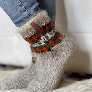 Op de afbeelding: Grijze wollen sokken met een patroonboord in oranje, groen en wit. Een klein bruin leren label is aan de boord bevestigd. De sokken worden gedragen met een blauwe jeans.