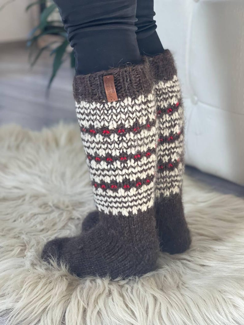 May include: Knee-high, hand-knitted socks in shades of brown, white, and red. The socks feature a patterned design with a brown cuff and foot. A small leather tag is attached to the cuff.