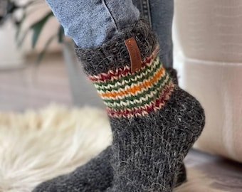 May include: Grey knitted socks with a striped cuff in red, green, orange, and white. A small brown leather tag is attached to the cuff. The socks are worn with blue jeans. The socks appear to be made of wool.