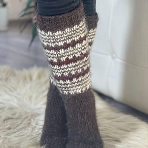 May include: A pair of brown and white knit leg warmers with a patterned design. The leg warmers have a dark brown cuff and foot, with a white and brown pattern and red accents. The leg warmers are made of a soft, textured material.