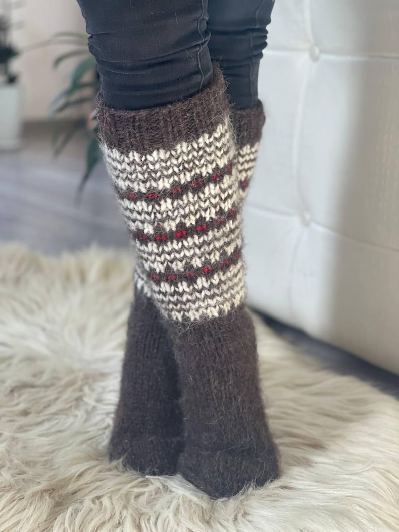 May include: A pair of brown and white knit leg warmers with a patterned design. The leg warmers have a dark brown cuff and foot, with a white and brown pattern and red accents. The leg warmers are made of a soft, textured material.