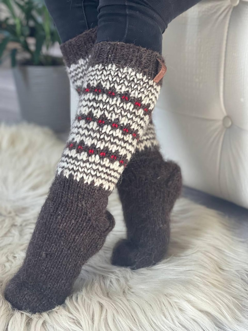 May include: Knee-high knit socks in brown, white, and red. The socks feature a patterned design on the upper portion and a solid brown color on the foot. A small leather tag is attached to the top of one sock.