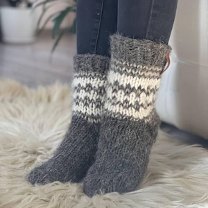May include: A pair of grey and white knitted socks with a geometric pattern. The socks are thick and appear warm, with a cuff detail. The socks are worn with dark jeans and rest on a fluffy, white rug. The socks are a cosy accessory.