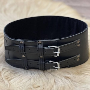 Op de afbeelding: Een zwarte leren riem met twee dubbele gespen en zilveren hardware. De riem is breed en heeft een gladde, gepolijste afwerking.