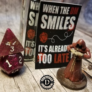 Op de afbeelding: Een rode en gouden glitter dobbelsteenset met een 1 en een 3 zichtbaar. Een miniatuurfiguur van een gekapte figuur in een rode mantel staat op een bruine basis. Een zwart-witte sticker met de tekst "When the DM Smiles It's Already Too Late" is op de achtergrond.