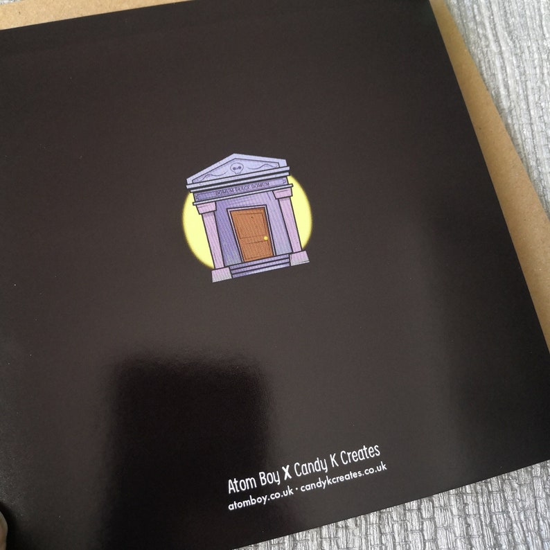Pu&ograve; includere: Carta nera con un'illustrazione di un cartone animato di un edificio viola con una porta e il testo "SPOOKY HOUSE GANG" sopra la porta. L'edificio &egrave; circondato da un cerchio giallo. Il testo "Atom Boy X Candy K Creates" e il sito web "atomboy.co.uk - candykkreates.co.uk" sono in fondo alla carta.