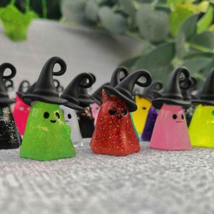 Puede incluir: Una colección de figuras de fantasmas coloridos y brillantes con sombreros de bruja negros. Los fantasmas son de varios colores, incluyendo verde, rojo, rosa y amarillo. Cada fantasma tiene una cara linda y simple. Las figuras son probablemente para Halloween.