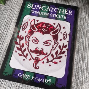 May include: A red glitter suncatcher sticker featuring a cartoon devil with horns and a moustache. The sticker is on a white background with the text "SUNCATCHER WINDOW STICKER" and "CANDY K CREATES".