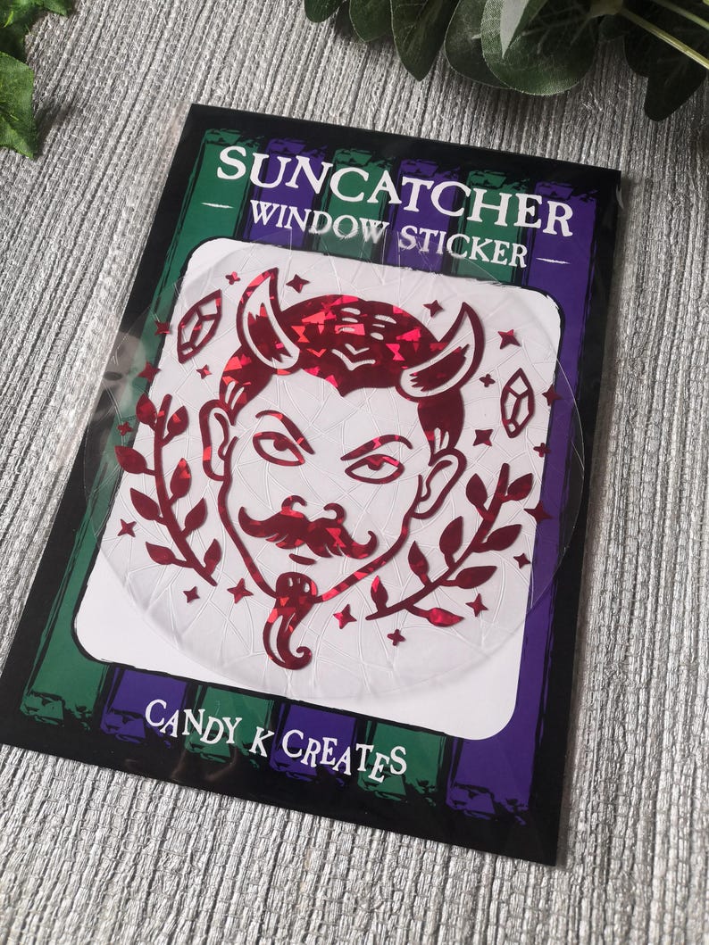 May include: A red glitter suncatcher sticker featuring a cartoon devil with horns and a moustache. The sticker is on a white background with the text "SUNCATCHER WINDOW STICKER" and "CANDY K CREATES".
