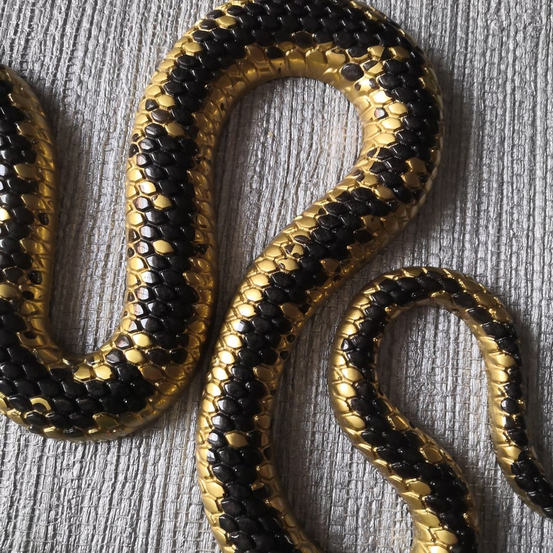 以下が含まれることがあります： 黒と金の蛇の彫刻で、質感のある鱗模様が施されています。蛇は8の字型に巻かれています。