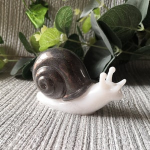 May include: A white snail figurine with a black and silver shell. The snail is sitting on a gray textured surface.