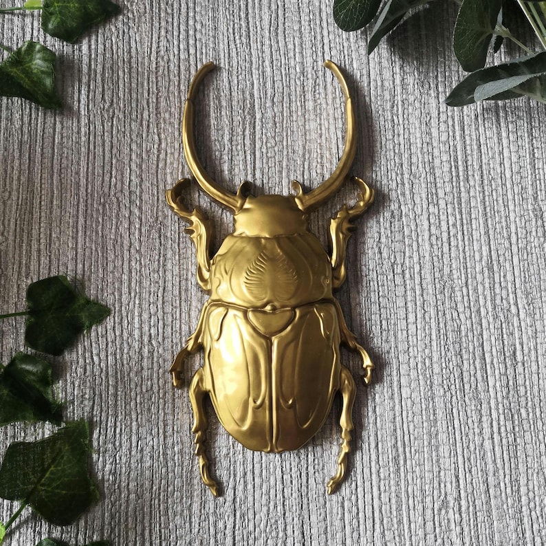 May include: A golden-colored, decorative beetle sculpture with large curved horns and detailed body segments. The sculpture is mounted on a textured, gray surface, with green leaves in the background.