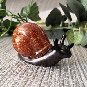 May include: A brown and black snail figurine with a glittery shell. The shell is a light brown color with gold glitter. The snail is made of resin and has a glossy finish.