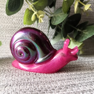 May include: A decorative snail figurine in shades of pink and purple. The snail's shell has a shimmering, iridescent finish, while the body is a solid, vibrant pink. The figurine is a whimsical decorative item.