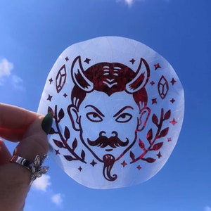May include: A red and white sticker featuring a devil face with horns and a moustache. The sticker is on a clear background with stars and leaves.