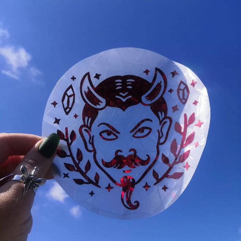 May include: A red and white sticker featuring a devil face design. The devil has horns, a moustache, and a goatee. The sticker is on a white background.