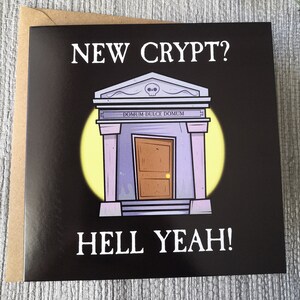 Pu&ograve; includere: Una carta nera con una scritta bianca che dice "NEW CRYPT? HELL YEAH!" La carta presenta un'illustrazione di un cartone animato di una cripta viola con una porta di legno e un teschio sopra la porta. La cripta ha un bagliore giallo dietro.