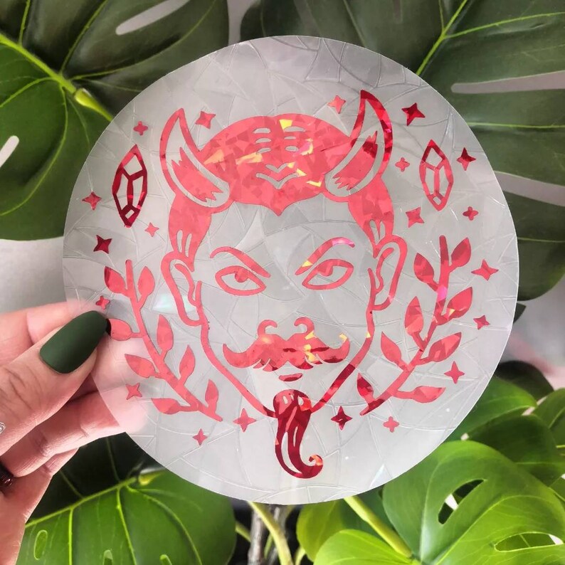 May include: A red holographic sticker featuring a devil face design. The devil has horns, a moustache, and a goatee. The sticker is surrounded by small stars and leaves.