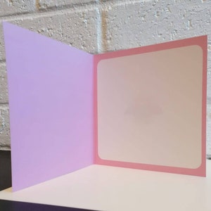 May include: A blank greeting card with a light pink inside and a lavender outside. The card is folded in half and is standing upright on a black and white chequered surface.