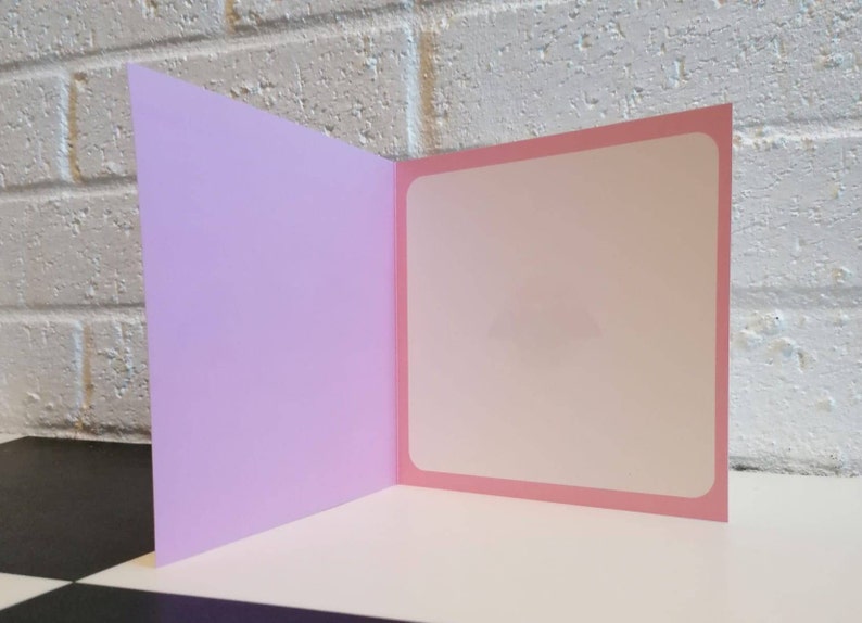 May include: A blank greeting card with a light pink inside and a lavender outside. The card is folded in half and is standing upright on a black and white chequered surface.