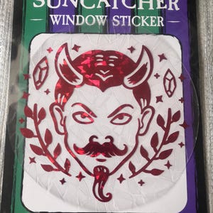 May include: A red holographic window sticker featuring a devil with horns, a moustache, and a goatee. The sticker is surrounded by stars and crystals. The text "SUNCATCHER WINDOW STICKER" and "CANDY K CREATES" are printed on the sticker.