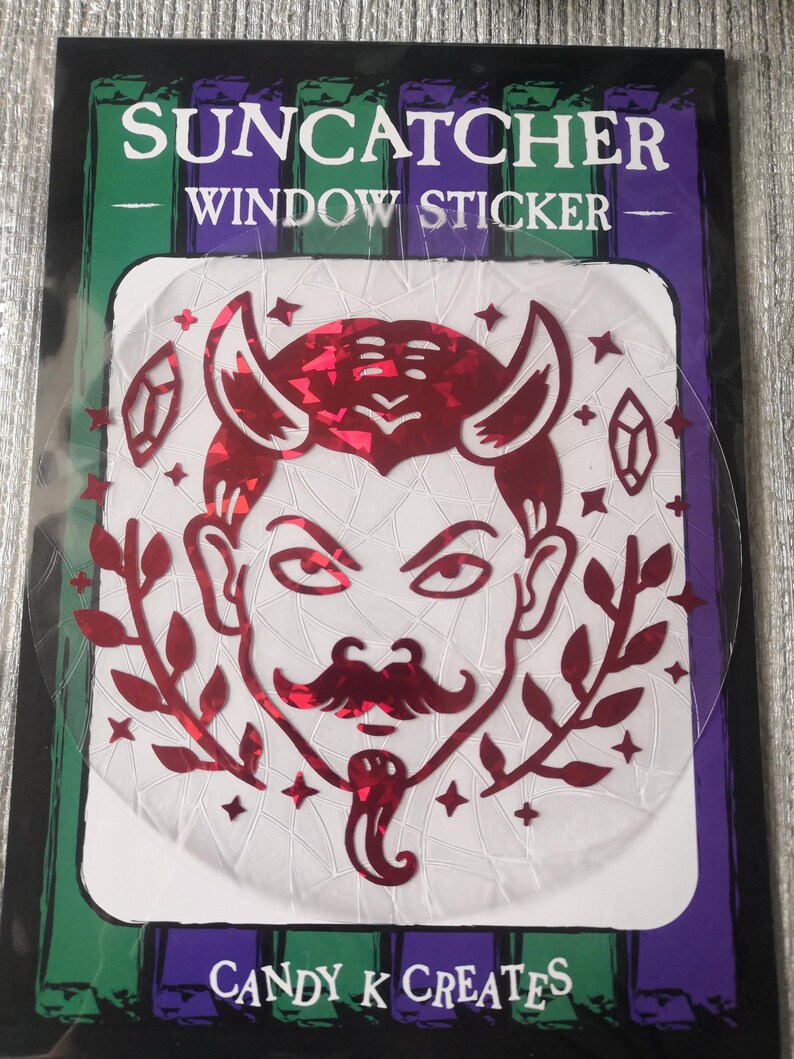 May include: A red holographic window sticker featuring a devil with horns, a moustache, and a goatee. The sticker is surrounded by stars and crystals. The text "SUNCATCHER WINDOW STICKER" and "CANDY K CREATES" are printed on the sticker.