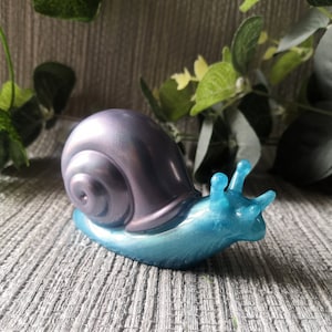 May include: A blue and purple iridescent snail figurine. The shell is a deep purple with a pearlescent finish. The body of the snail is a bright blue with a glittery finish.