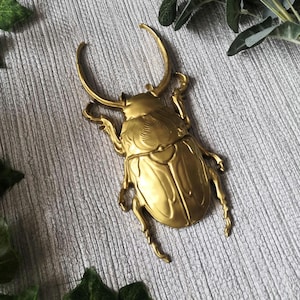 May include: A detailed, golden-colored beetle sculpture with large curved horns and intricate wing detailing. The beetle is positioned on a textured, gray surface, showcasing its metallic sheen. The sculpture's legs are also visible.