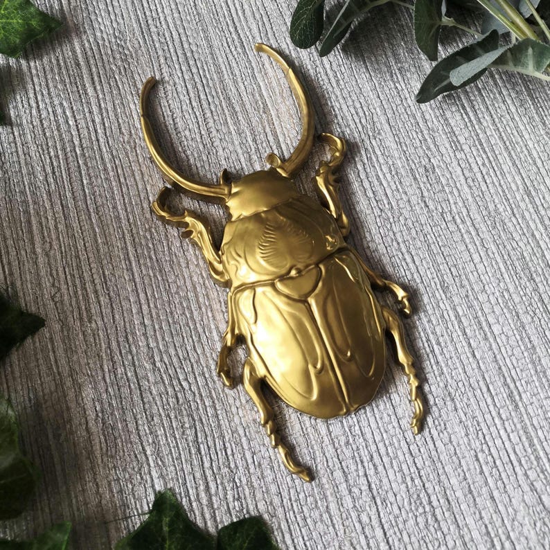 May include: A detailed, golden-colored beetle sculpture with large curved horns and intricate wing detailing. The beetle is positioned on a textured, gray surface, showcasing its metallic sheen. The sculpture's legs are also visible.