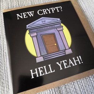 Pu&ograve; includere: Un biglietto di auguri in bianco e nero con un'illustrazione a fumetti di una cripta con il testo "NEW CRYPT? HELL YEAH!" e la frase latina "DOMUM DULCE DOMUM" sopra la porta.