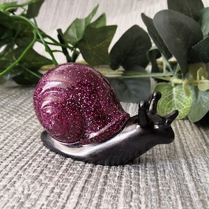May include: A black and purple snail figurine with a glittery purple shell. The snail is made of resin and has a glossy finish.