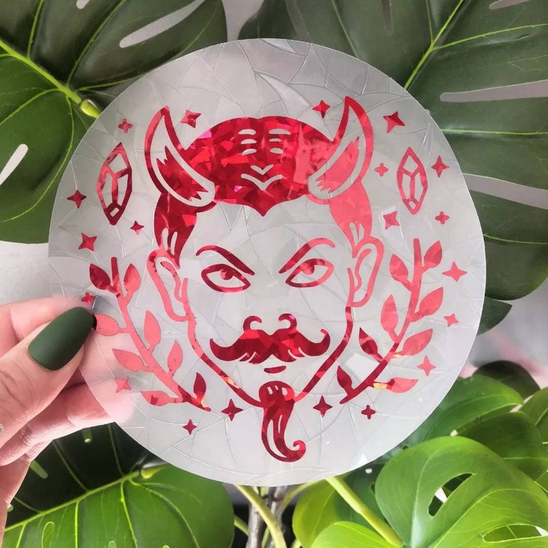 May include: A red glitter vinyl sticker of a devil with horns, a moustache, and a goatee. The sticker is on a clear background with a cracked glass effect. The sticker is surrounded by small stars and leaves.