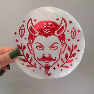 May include: Red holographic sticker featuring a devil face design. The devil has horns, a moustache, and a goatee. The sticker is surrounded by stars and leaves.