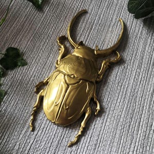 May include: A golden-colored, decorative beetle sculpture. The beetle has large, curved horns and detailed wings and legs. The sculpture is made of metal and has a shiny finish. The beetle is a decorative item.