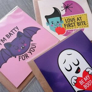 May include: Three pink and purple greeting cards with humorous Halloween-themed illustrations and text. The first card features a purple bat with the text "I'm batty for you!" The second card features a cartoon vampire with the text "Love at first bite." The third card features a ghost with the text "Be my boo?"