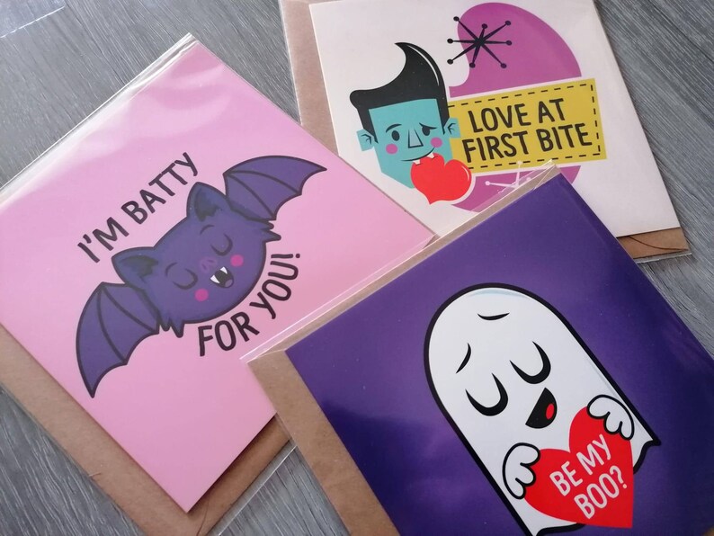 May include: Three pink and purple greeting cards with humorous Halloween-themed illustrations and text. The first card features a purple bat with the text "I'm batty for you!" The second card features a cartoon vampire with the text "Love at first bite." The third card features a ghost with the text "Be my boo?"