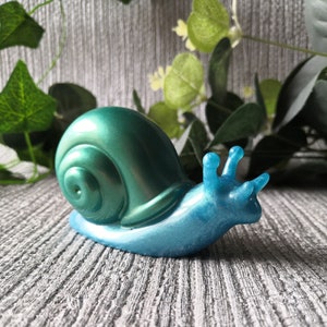 May include: A teal blue and green snail figurine with a shiny, iridescent finish. The snail is in a crawling position with its shell facing the left side of the image.