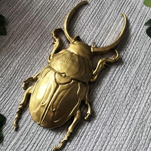 May include: A detailed, golden-colored beetle sculpture with large curved horns and intricate detailing on its body. The sculpture is made of metal and has a shiny finish, giving it a luxurious appearance. The beetle is a decorative item.