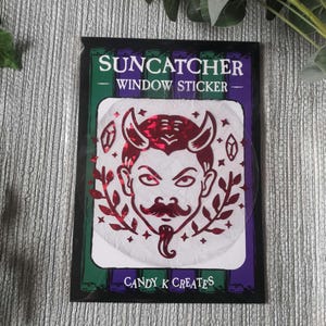 May include: Red and white window sticker featuring a devil face design. The sticker reads "Suncatcher Window Sticker" and "Candy K Creates".