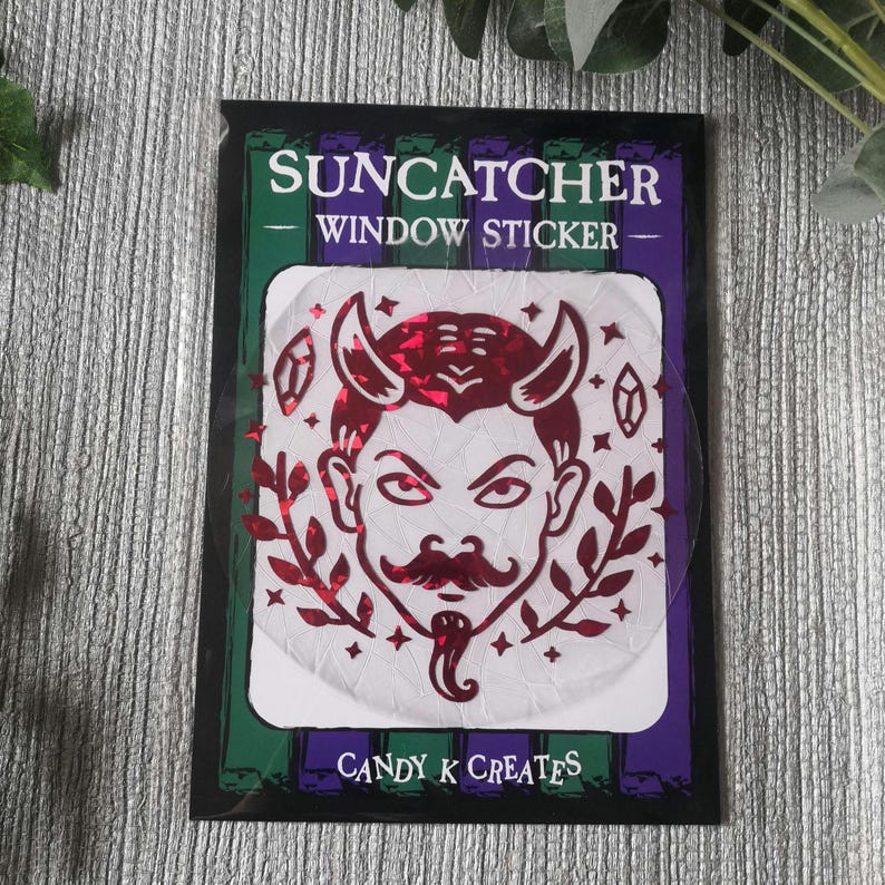 May include: Red and white window sticker featuring a devil face design. The sticker reads "Suncatcher Window Sticker" and "Candy K Creates".