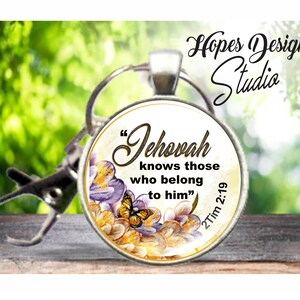 Peut inclure: Un porte-clés rond en verre avec un fond blanc et un motif floral. Le texte "Jéhovah connaît ceux qui lui appartiennent" est imprimé en noir avec un contour doré. Le verset biblique "2 Tim 2:19" est imprimé en noir sur le côté droit du porte-clés.