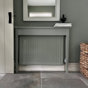 May include: A grey painted radiator cover with a white base and a white trim around the top. The cover is sitting on a grey tiled floor. A book is sitting on top of the cover.