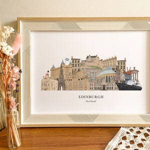 May include: A framed print of a colorful illustration of the Edinburgh skyline, featuring iconic landmarks like Edinburgh Castle and the Scottish Parliament. The print is in a white frame with a wood-like finish and has the text "EDINBURGH Scotland" at the bottom.