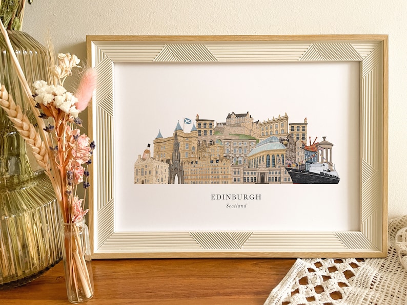 May include: A framed print of a colorful illustration of the Edinburgh skyline, featuring iconic landmarks like Edinburgh Castle and the Scottish Parliament. The print is in a white frame with a wood-like finish and has the text "EDINBURGH Scotland" at the bottom.